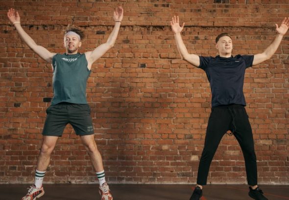 Two men performing jumping jacks in a gym setting, showcasing fitness and energy.