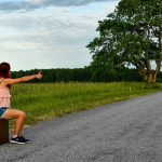 A woman sits on a suitcase, hitchhiking along a peaceful country road in Kolga, Estonia.
