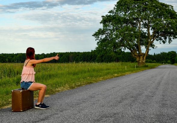 A woman sits on a suitcase, hitchhiking along a peaceful country road in Kolga, Estonia.