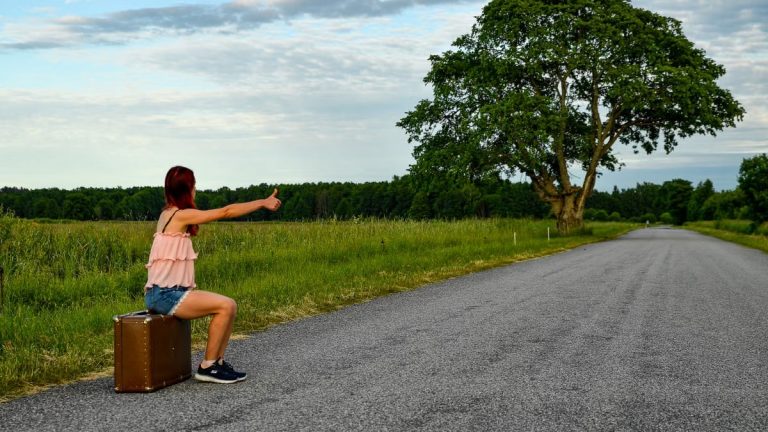 A woman sits on a suitcase, hitchhiking along a peaceful country road in Kolga, Estonia.